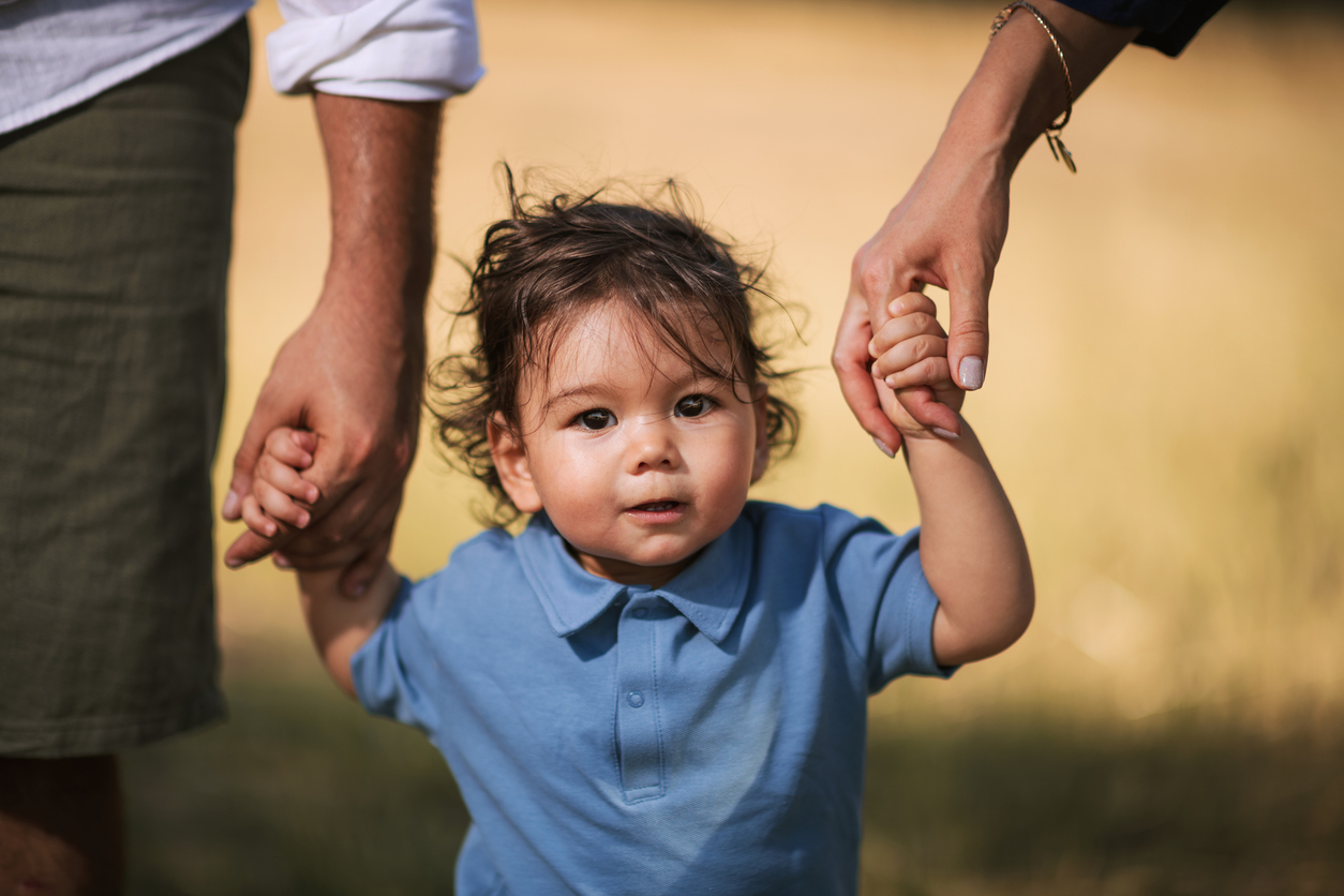 A one-year-old boy walking on the grass, holding his parents' hands, wearing a blue shirt and blue denim shorts.