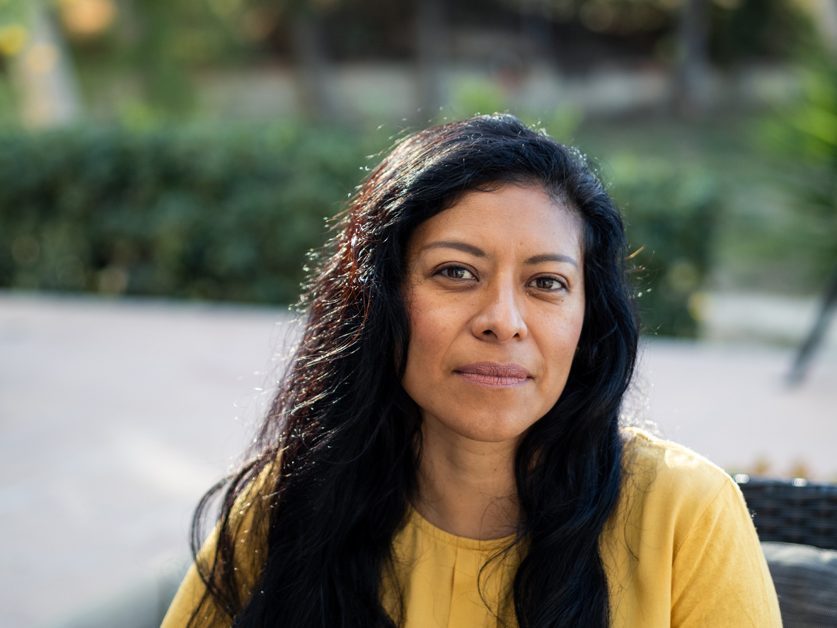 A Mexican woman with long dark hair sits outdoors, wearing a bright yellow blouse, exuding confidence and warmth. Sunlight reflects off her face as she relaxes in a serene garden setting.