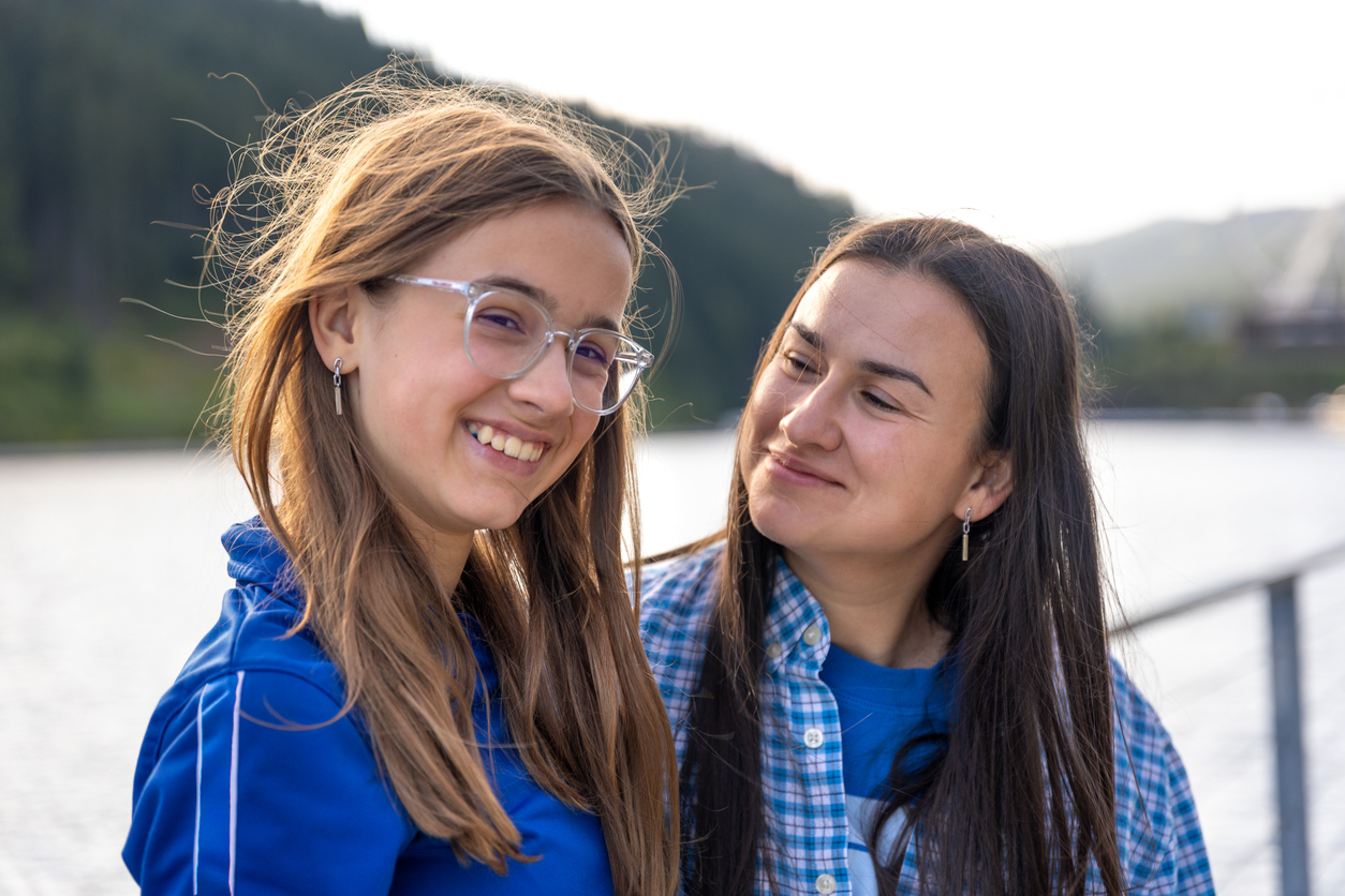 Half length portrait on the pier. Daughter with clear glasses smiles while mom looks with tenderness, everyday casual and warm light.