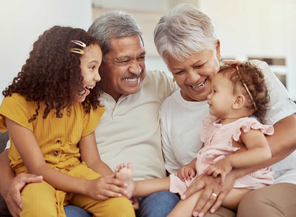 Mixed race grandparents enjoying weekend with granddaughters in home living room. Adorable smiling hispanic girls bonding with grandmother and grandfather. Happy seniors and children sitting together