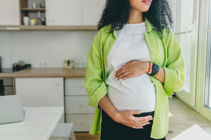 No face picture of pregnant african American woman with long curly hair looking through window standing in kitchen, rubbing her big cute belly, expecting baby boy to be born in few month