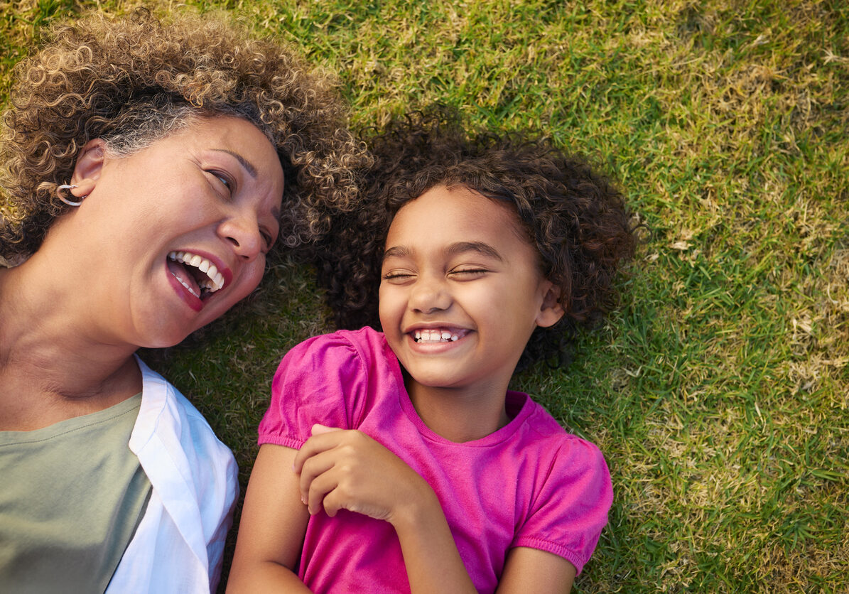Looking Down On Grandmother With Granddaughter Lying Outdoors On Grass Looking Up At Camera