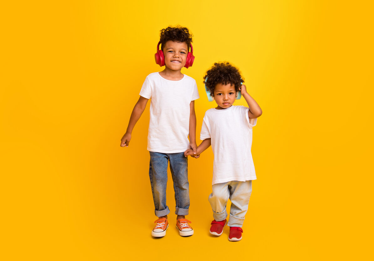 Full length photo of cheerful positive small boys wear white t-shirt walking hands listening music headphones isolated yellow color background.